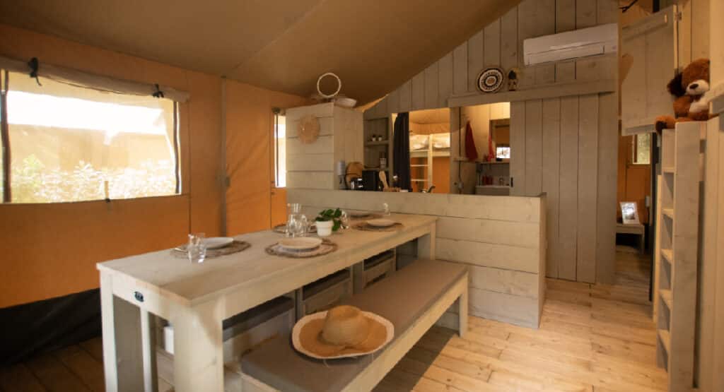 Interior of a safari tent at Yelloh! Village Sant Miquel campsite. The tent features a rustic dining area with a wooden table and benches, set with plates and glasses. The space is well-lit with natural light from large windows. In the background, a compact kitchen and a bunk bed area can be seen, highlighting the cozy and well-equipped accommodations.