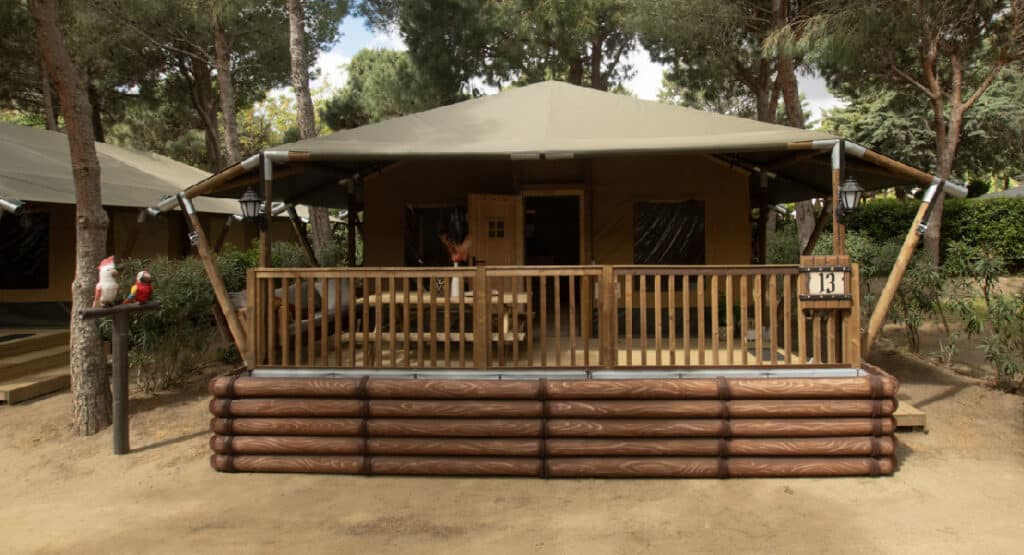 Front view of a safari tent at Yelloh! Village Sant Miquel campsite. The tent is set on a wooden platform with log-style railings and features a spacious porch with a dining table. The area is shaded by tall pine trees, creating a cozy and natural atmosphere. Decorative ceramic parrots are perched on a stand to the side, adding to the charming and playful ambiance of the campsite.